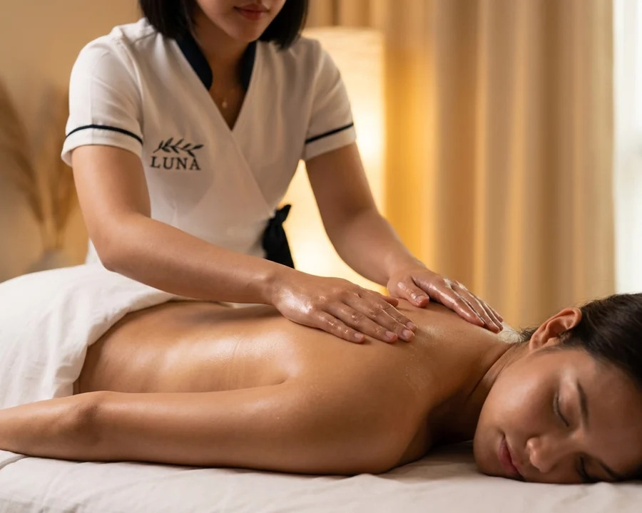 A Luna therapist working warm coconut oil into the back and shoulders of a relaxed client on fresh white sheets during an at-home coconut oil massage in Bangkok